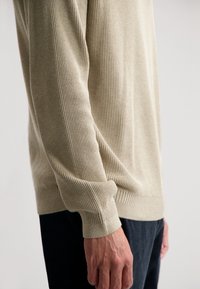 Beige gerippter Pullover mit langen Ärmeln, der über ein strukturiertes Strickmuster und gerippte Bündchen verfügt. Das Modell trägt dunkle Hosen und zeigt eine lockere Passform.