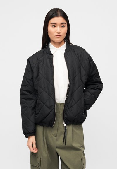 Μαύρο μπουφάν bomber με καπιτονέ ύφασμα, με ριμπ μανσέτες και ζώνη στη μέση, κλείσιμο με φερμουάρ και εργονομικό σχεδιασμό. Φορεμένο πάνω από λευκό πουκάμισο με γιακά.