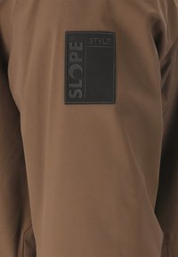 Brun yderbeklædning med en rektangulær sort lap, der har teksten "SLOPE" og "STYLE". Stoffet ser glat ud med en mat finish.