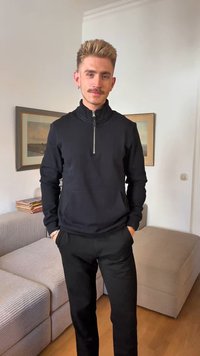 Black zip-up sweatshirt with long sleeves, featuring a high collar and front pockets, paired with black pants on a light wood floor.