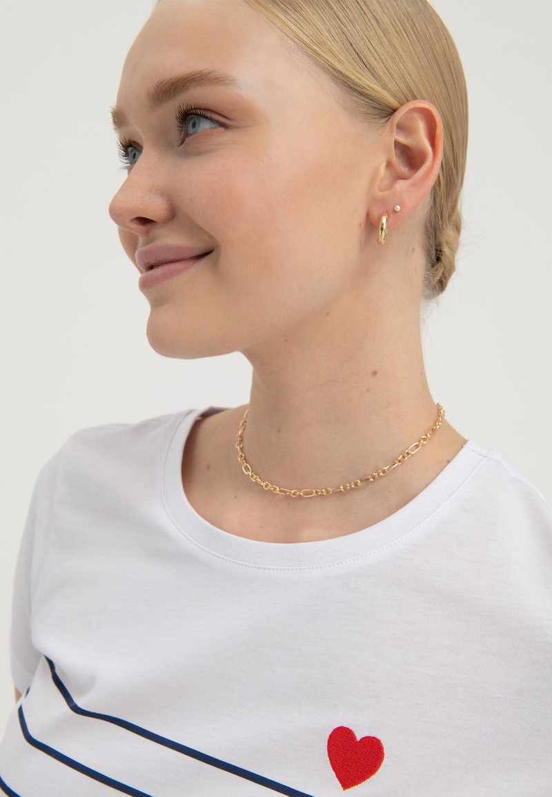 T-shirt bianco con righe blu, dettaglio a forma di cuore rosso; collana in catena dorata; capelli raccolti in una treccia, occhi azzurri, trucco naturale.
