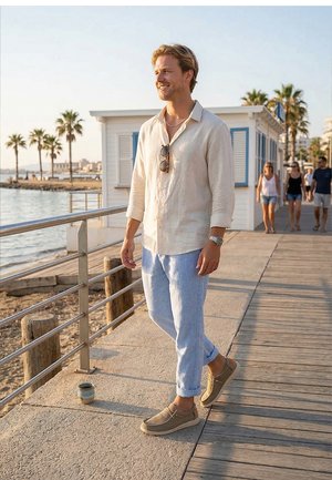 Hombre con camisa beige y pantalones azul claro caminando por un paseo marítimo con palmeras y personas al fondo al atardecer.