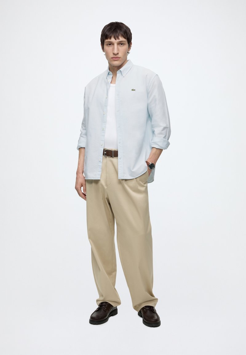 Giovane uomo che indossa una camicia azzurro chiaro con bottoni sopra una maglietta bianca, pantaloni beige, cintura marrone, scarpe scure e orologio da polso, in piedi con la mano in tasca.