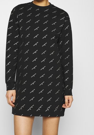 Osoba nosící černé šaty s dlouhým rukávem, celé pokryté malými bílými šikmými logy "Calvin Klein Jeans".