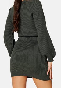 Femme portant une robe pull en tricot côtelé vert foncé avec des manches longues bouffantes, vue de dos avec de longs cheveux bruns tombant.
