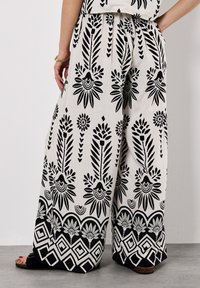 Pantalons larges en tissu léger avec des motifs floraux noirs et géométriques sur fond beige. Taille élastique et poches latérales.