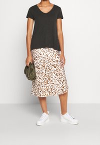 Person trägt ein schwarzes V-Ausschnitt-T-Shirt, einen weißen Midirock mit braunen Flecken, weiße Sneaker und hält eine kleine olivgrüne Handtasche.