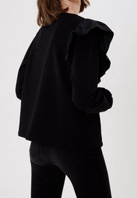 Mujer vestida con una blusa negra de manga larga con detalle de volantes en los hombros y pantalones negros ajustados, de pie contra un fondo blanco liso.