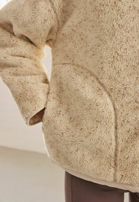 Flauschige beige Fleecejacke mit strukturiertem Obermaterial, lockerer Passform, Seitentaschen und glattem Saum mit leichter Stoffauskleidung.