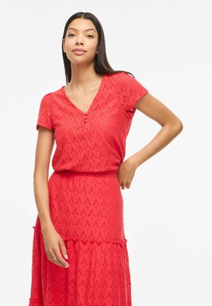 Femeie purtând un top roșu cu decolteu în V și mâneci scurte cu model ochiuri și o fustă asortată, stând cu o mână pe șold.