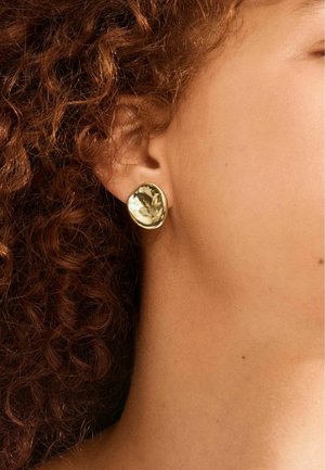 Capelli ricci castano ramato e un orecchino a perno irregolare dorato sull'orecchio di una persona con pelle di colore marrone chiaro.