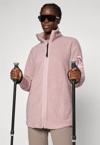 Helle pinke Fleecejacke mit hohem Kragen, Frontreißverschluss, strukturiertem Stoff und kombiniert mit schwarzen Wanderstöcken. Knopfdetails an den Ärmeln.