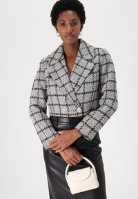 Kuttet dobbeltbrystet jakke i grå og svart rutete tweed med store slag og to dekorative knapper. Modellen holder en liten hvit veske.