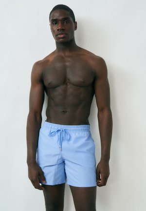 Skjorteløs mand står mod en hvid væg iført lyseblå badebukser med snor, vendt fremad med afslappet kropsholdning.