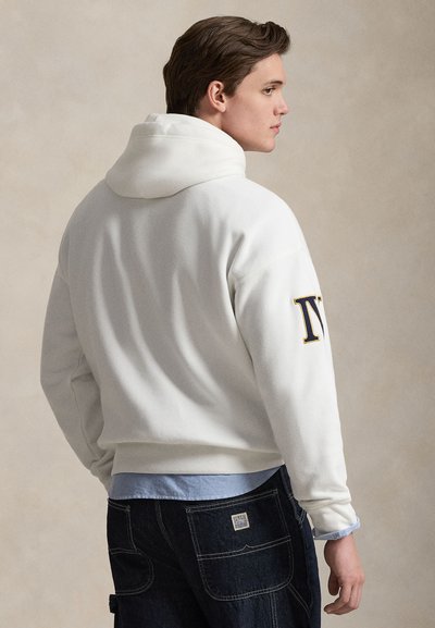 Młody mężczyzna stojący bokiem, ubrany w biały kaptur z napisem "NY" na prawym rękawie oraz ciemne dżinsy, na jasnobeżowym, gładkim tle.