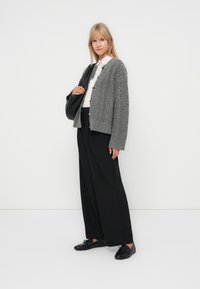 Grijze kabelgebreide cardigan met knoopsluiting, oversized pasvorm, gedragen over een wit shirt, gecombineerd met zwarte wijde broek en zwarte schoenen.