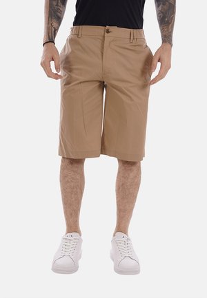 Uomo che indossa pantaloncini beige fino al ginocchio, camicia nera, scarpe da ginnastica bianche e tatuaggi visibili sugli avambracci, in piedi con le mani rilassate lungo i fianchi.