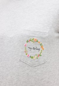 Τσεπάκι από ανοιχτό γκρι ύφασμα με κεντητό στεφάνι από λουλούδια σε ροζ, πορτοκαλί και πράσινο, που περιβάλλει το κείμενο «Hey darling.»