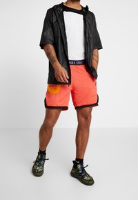 Veste imperméable noire, t-shirt à col ras du cou blanc, shorts orange vif avec poches jaunes et chaussures de sport multicolores.