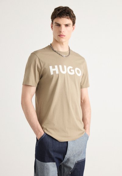 Бежова памучна тениска с голям бял текст "HUGO". Моделът я носи с дънкови панталони с пачwork. Къси ръкави, дизайн с кръгло деколте.
