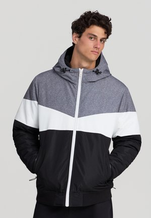 Giovane uomo che indossa una giacca color block con cappuccio grigio, striscia centrale bianca e parte inferiore nera, in piedi con le mani in tasca.