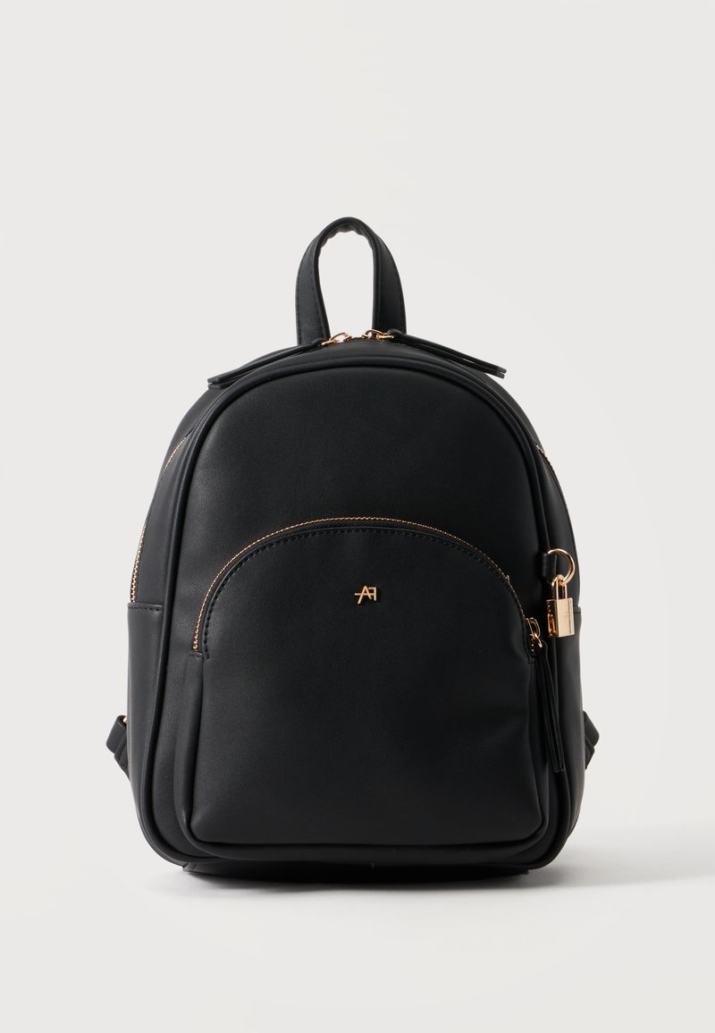 Petit sac à dos en cuir noir avec des détails de fermeture éclair dorés et une poche avant avec un petit logo doré et un charm cadenas doré sur le côté.