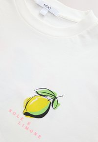 Bílé tričko s grafikou žlutého citronu a zelených listů a růžovým textem s nápisem "SOLE E LIMONE" pod citronem.