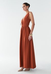 Rūdžių oranžinės spalvos maksi suknelė su gilia V formos iškirpte, prigludusia juosmens dalimi ir besileidžiančia suknele. Pasižymi raišteliu priekyje ir lygiu paviršiumi.