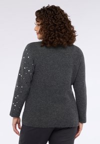 Donkergrijze trui met een geribbelde textuur, voorzien van lange mouwen met verspreide zilveren pailletten langs één arm. Casual pasvorm, afgeronde zoom.