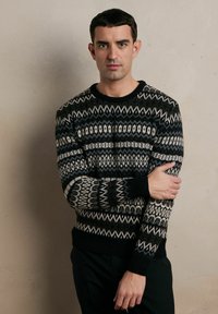 Czarny sweter z kremowym wzorem, wykonany na drutach, z geometrycznymi motywami, okrągłym dekoltem i ściągaczami. Model stoi na tle w neutralnych kolorach.