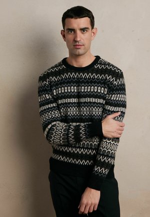 Czarny sweter z kremowym wzorem, wykonany na drutach, z geometrycznymi motywami, okrągłym dekoltem i ściągaczami. Model stoi na tle w neutralnych kolorach.