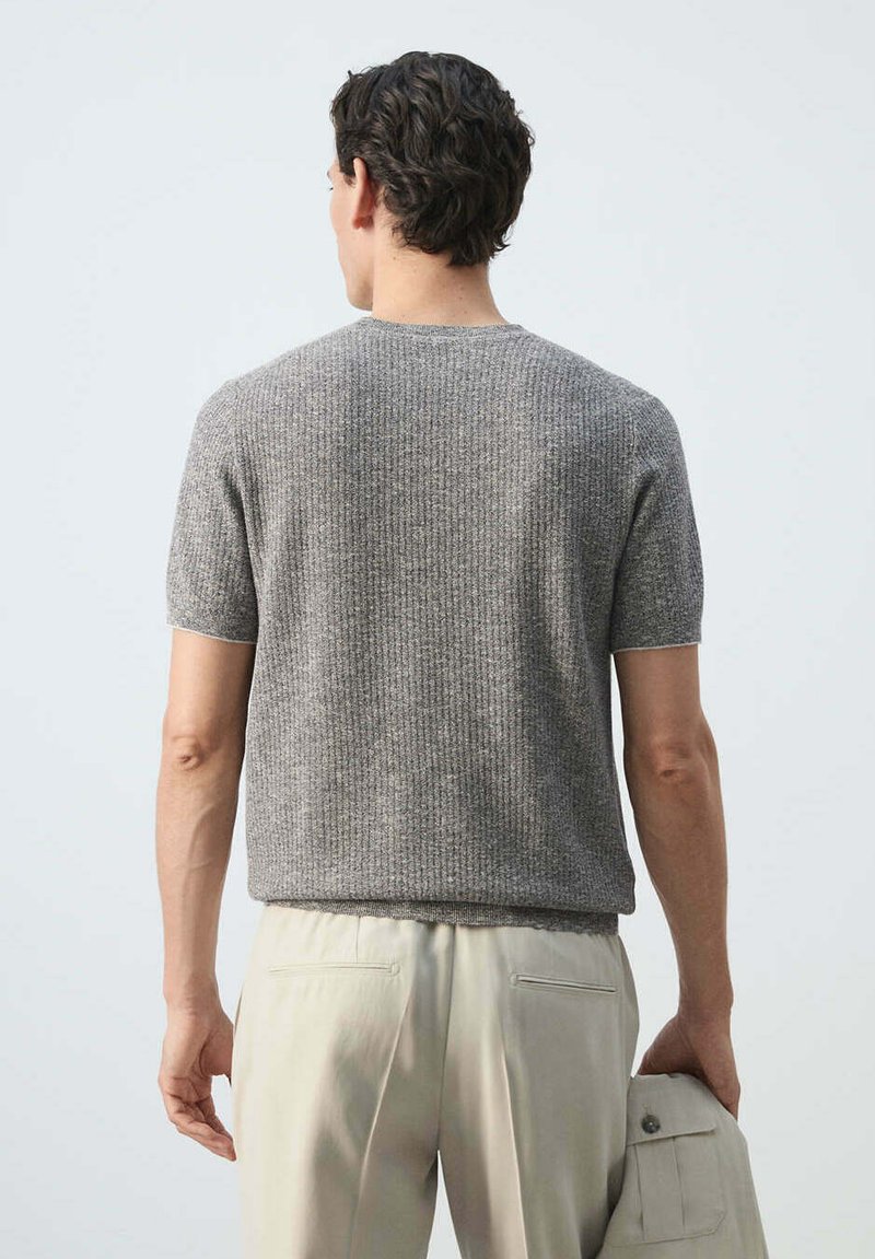 Homme aux cheveux foncés ondulés portant un pull en maille gris à manches courtes et un pantalon beige clair, tenant une veste beige assortie dans la main droite.