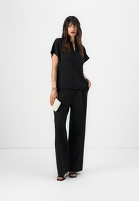 Black short-sleeve top with a V-neck, paired with wide-leg black trousers. Model holds a white textured clutch, wearing black sandals.