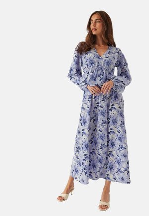 Kvinde iført en lang, blå og hvid blomsterkjole med puffede, lange ærmer, stående med hænderne foldet og iført beige åbne hæle.