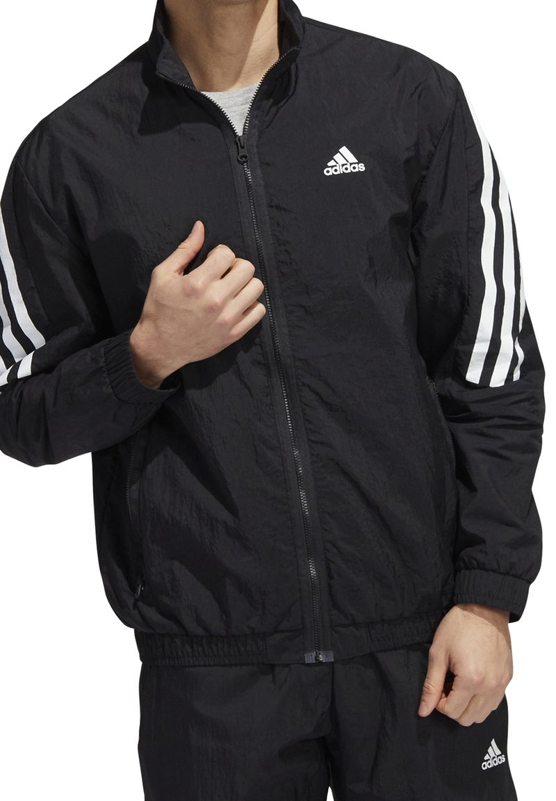 Svart zip-up jacka i strukturerat tyg. Har vitt tre-stripes mönster på ärmarna och en broderad logo på bröstet. Elastiska muddar.