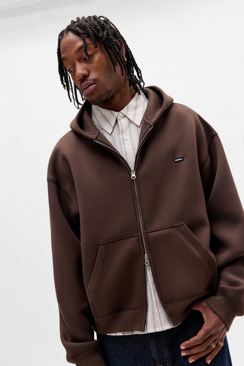 Felpa con zip marrone realizzata con tessuto morbido, con cappuccio regolabile con coulisse, due tasche frontali e una piccola etichetta con logo sul petto.