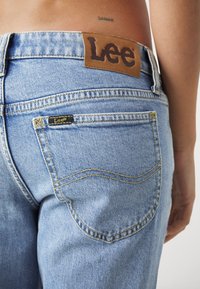Jean en denim bleu clair avec étiquette en cuir de la marque Lee sur la poche arrière, porté par une personne avec un petit tatouage "SANAR" au-dessus de la ceinture.