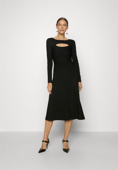 Μαύρο φόρεμα με μακριά μανίκια, τετράγωνη λαιμόκοψη και κοψίματα. Αμπιγιέ σιλουέτα, μαλακή υφή και εφαρμοστό σώμα. Φοράει μαύρες γόβες.