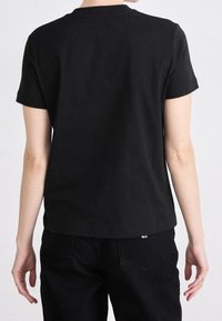 Osoba w jednolitym czarnym t-shircie z krótkim rękawem i czarnych spodniach, pokazana od tyłu na neutralnym tle.