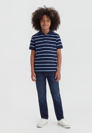 Παιδί που στέκεται με το πρόσωπο προς τα εμπρός, φοράει μπλε σκούρο πόλο πουκάμισο με ανοιχτό μπλε ρίγες, σκούρο τζιν και λευκά αθλητικά παπούτσια σε λευκό φόντο.