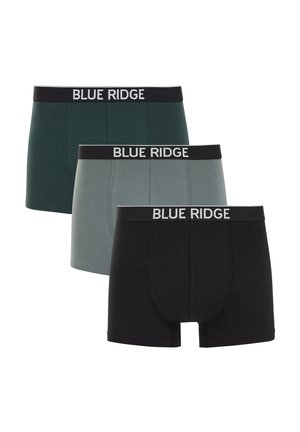 Mężczyzna bez koszulki z krótkimi kręconymi włosami, ubrany w ciemnozielone bokserki Blue Ridge, stoi na tle jednolitego beżowego tła.