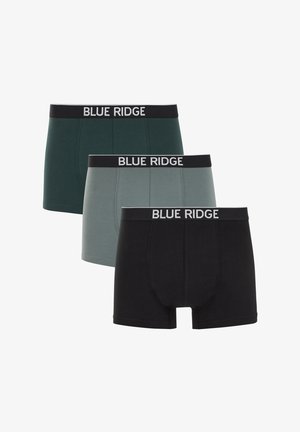 Särgi seljast võtnud mees, kellel on lühikesed lokkis juuksed ja kes kannab tumerohelisi Blue Ridge bokserkorsette, seisab tavalise beežika tausta ees.