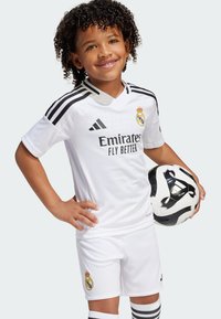 Bijela nogometna oprema s crnim detaljima, s logom Real Madrida. Kratki rukavi, kratke hlače i crno-bijela nogometna lopta u ruci.