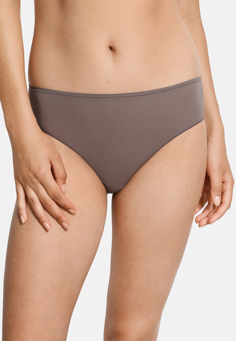 Frau trägt ein schlichtes braunes Bikini-Unterwäsche-Set vor einem hellgrauen Hintergrund, zeigt dabei Bauch und Oberschenkel.