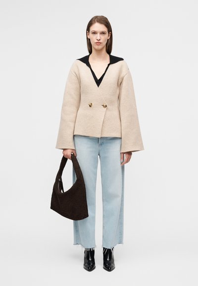 Μπεζ διπλόμπουτο μάλλινο σακάκι με μαύρο γιακά, συνδυασμένο με ανοιχτό μπλε τζιν με φαρδιά μπατζάκια, κρατώντας μια σκούρα καφέ τσάντα με υφή και μαύρα μποτάκια με τακούνι.