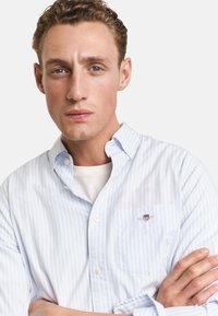 Hellblau-weiß gestreiftes Hemd mit Knopfleiste und kleinem Brusttaschen-Logo. Der Stoff wirkt glatt und leicht strukturiert.