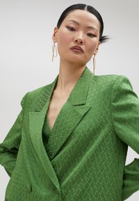 Blazer verde con un patrón, de escote en V profundo y solapas estructuradas. Textura suave y corte a medida, complementado con pendientes geométricos dorados.