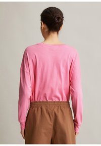Frau mit dunklem Haar in einem Dutt, die ein rosa Langarmshirt und braune Hose mit elastischem Bund trägt, von hinten vor einem einfarbigen Hintergrund gezeigt.