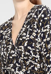 Blommig klänning med marinblå bakgrund, vitt och guldigt blommönster, v-ringning och lättviktsmaterial med en mjuk textur.