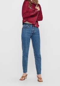 Femme portant une veste zippée rouge, un jean taille haute bleu et des sandales à talons ouvertes marron, debout devant un fond uni.